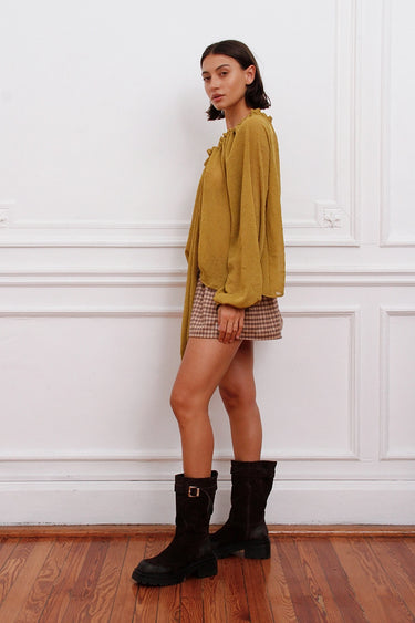 Blusa Klen Sofia Sarkany mostaza en modelo, estilo oversize, combinada con minifalda a cuadros y botas negras.