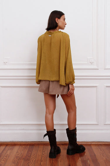 Blusa Klen mostaza Sofia Sarkany, mujer de espaldas luciendo blusa manga larga y pollera cuadrillé, look casual chic.
