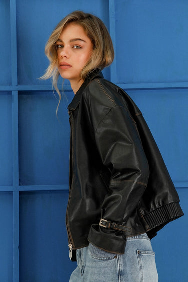Modelo con campera Sofia Sarkany Traf negra de cuero sintético, combinada con jeans y fondo azul vibrante.